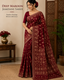 A woman models a deep maroon jamdani saree with intricate gold floral patterns, standing in a styled indoor setting with a wooden screen, potted plant, and a side table.